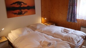Una cama bien hecha en una habitación con paneles de madera, una mesita de noche con una lámpara y un cuadro de un árbol junto al agua.