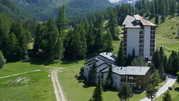 Una cabaña de montaña con un edificio grande y una extensión más pequeña, rodeada de un bosque denso y montañas nevadas.