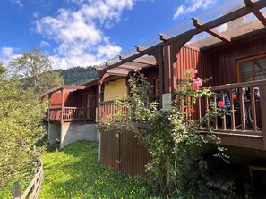 En træchalet med balkon, omgivet af frodig vegetation og et bjergrigt landskab.