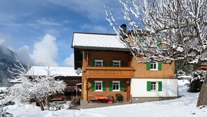 Un chalet à deux étages doté d’un balcon, entouré d’arbres couverts de neige et avec une chaîne de montagnes en toile de fond.