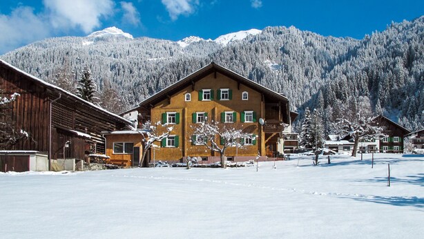 Un grand bâtiment à deux étages, de style architectural alpin traditionnel, entouré d’arbres et de montagnes couverts de neige.
