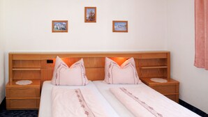 Une chambre d’hôtel avec une tête de lit en bois, deux tables de chevet, un lit recouvert d’une courtepointe blanche et agrémenté d’oreillers assortis, et trois tableaux encadrés accrochés au mur.