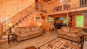 A spacious wooden cabin with a central living area featuring two sofas, a dining table, and a staircase leading to an upper level. - Cozy secluded cabin with firepit! - Absolutely Secluded (Gatlinburg)