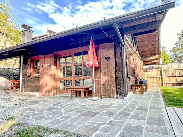 A rustic wooden cabin with a stone patio, a covered outdoor seating area, and a fenced backyard.