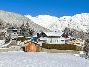 A snow-covered mountain landscape with a modern house featuring a sloped roof, large windows, and a wooden barn in the foreground.