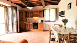A kitchen with wooden cabinets, a dining table with chairs, a TV mounted on the wall, and a window with curtains.
