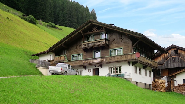 Un chalet traditionnel en bois, doté d’un balcon, avec un camion garé devant et une pile de bois de chauffage sur la droite.