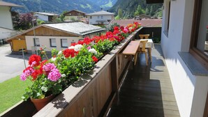 Un balcon avec des balustrades en bois, une rangée de fleurs aux couleurs vives et, au loin, une vue sur des maisons et des montagnes.