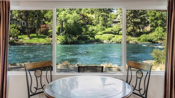 A room with a large window overlooking a river, two chairs, and a round table.