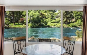 Een kamer met een groot raam met uitzicht op een rivier, twee stoelen en een ronde tafel.
