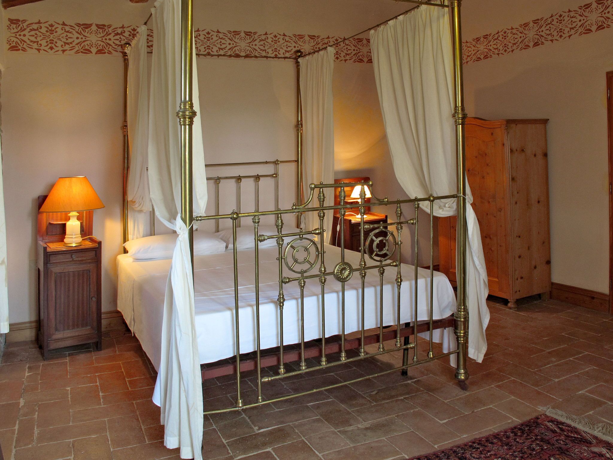 Una cama con dosel sostenida por cuatro columnas, con estructura de latón, ropa de cama blanca y cortinas colgantes.