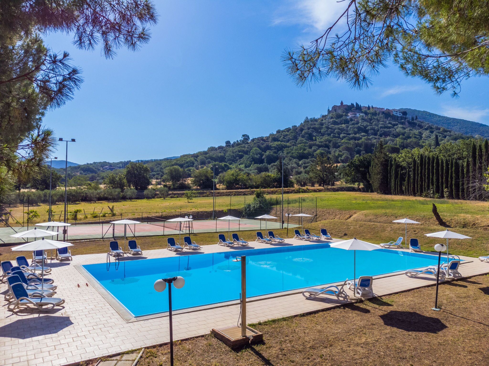 A swimming pool with lounge chairs and umbrellas, surrounded by a grassy area and trees, with a hill and building in the background.