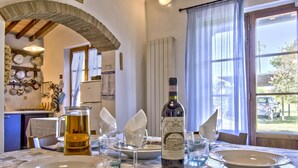 A dining area with a table set for two, a bottle of Chianti wine, and a pitcher of beer. The room features a rustic stone archway, a window with a view of the outside, and a kitchen area in the background. - Oliveta by Interhome (Cecina)