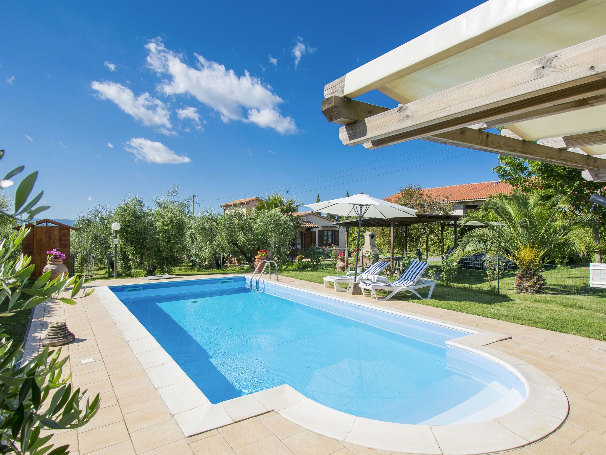 Una piscina rettangolare con superficie d'acqua cristallina e blu, circondata da un solarium piastrellato. Sono presenti lettini e ombrelloni sotto una struttura a pergola.