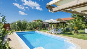 A rectangular swimming pool with a clear blue water surface, surrounded by a tiled deck. There are lounge chairs and umbrellas under a pergola structure. - Oliveta by Interhome (Cecina)