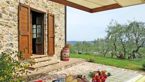 Un edificio de piedra con puerta y contraventanas de madera, una mesa con comida y bebidas, y vistas a un paisaje verde con árboles.