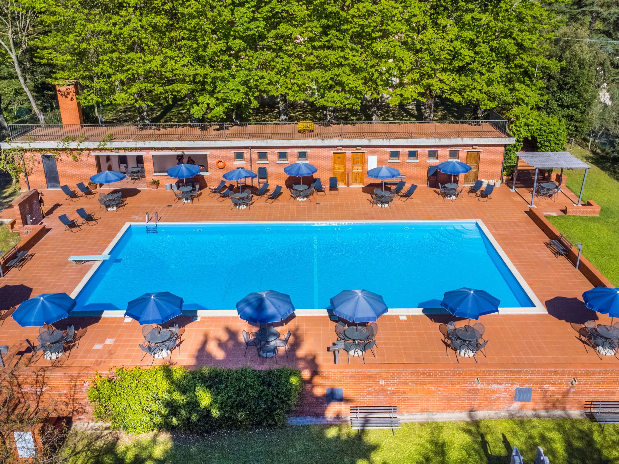 A rectangular swimming pool with lounge chairs and umbrellas around it, a brick building with multiple doors, and a covered seating area.