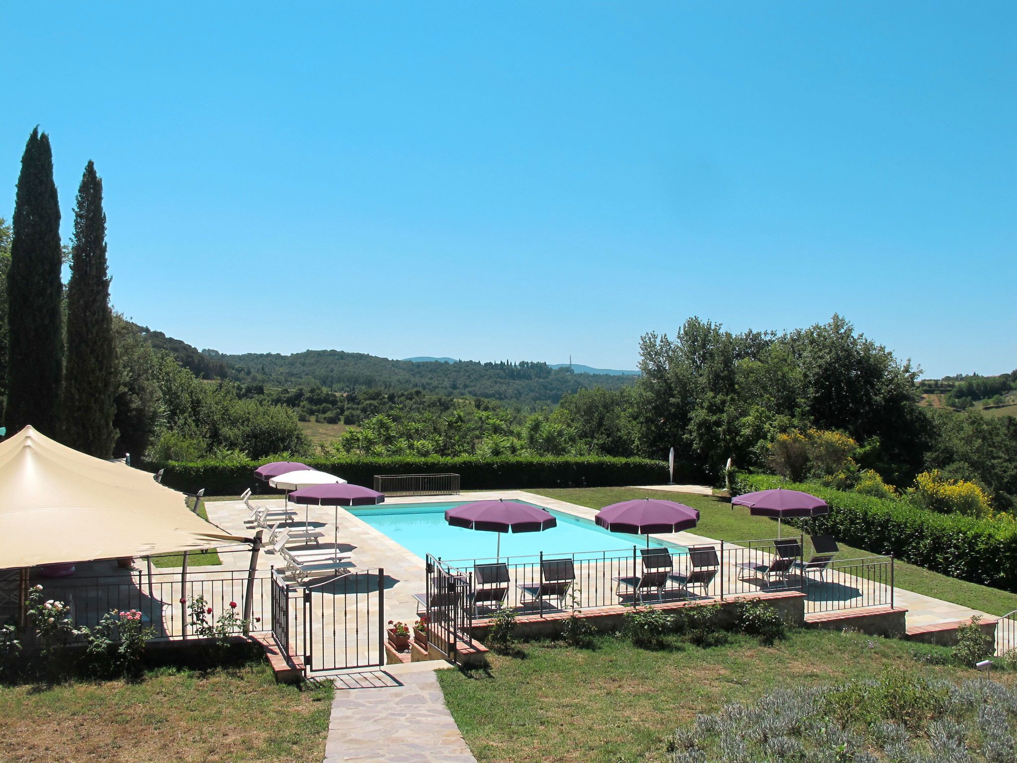 A pool area with multiple sun loungers and umbrellas, surrounded by a landscaped garden and a view of rolling hills.