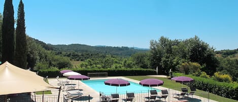 Un espace piscine doté de nombreuses chaises longues et de parasols, entouré d’un jardin aménagé et offrant une vue sur des collines verdoyantes.