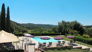 A pool area with multiple sun loungers and umbrellas, surrounded by a landscaped garden and a view of rolling hills.