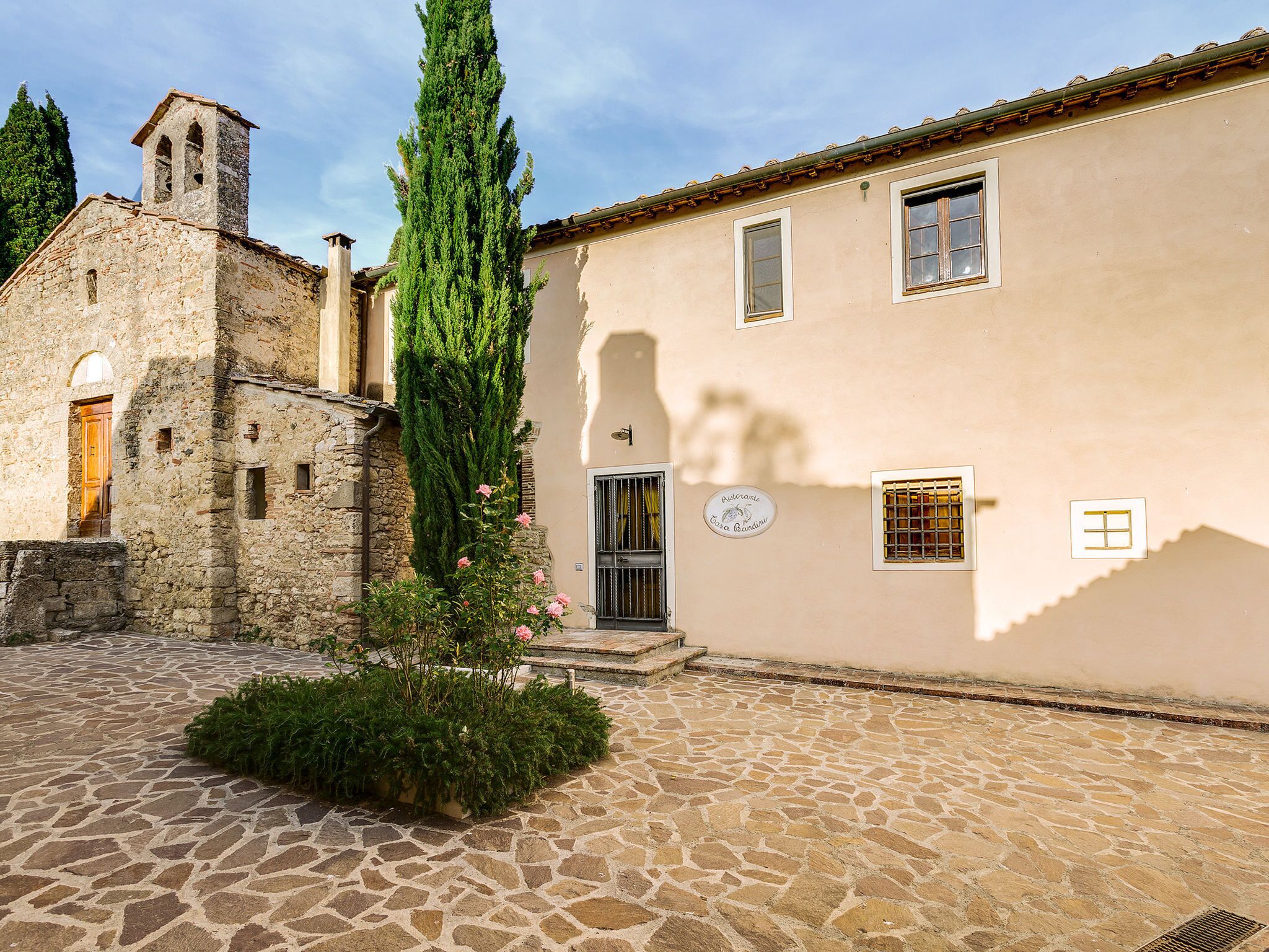 Un cortile con pavimento in pietra, un alto cipresso e un edificio con un campanile.