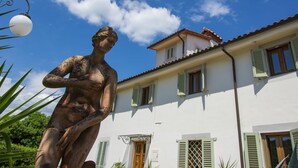 Une statue d’une femme, placée dans un jardin, avec un bâtiment blanc en arrière-plan.