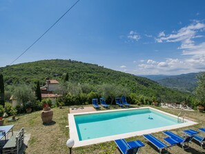 A rectangular swimming pool with blue lounge chairs, a wooden deck, and a view of a green hillside.