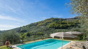 A swimming pool with lounge chairs and umbrellas, surrounded by a wooden deck and overlooking a hillside with trees and buildings. - Francesco by Interhome (Montecatini Terme)
