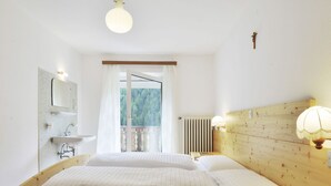 Une chambre à coucher avec un lit en bois, une table de chevet, une lampe de chevet, une fenêtre donnant sur des arbres, et une salle de bain avec un lavabo et un miroir.