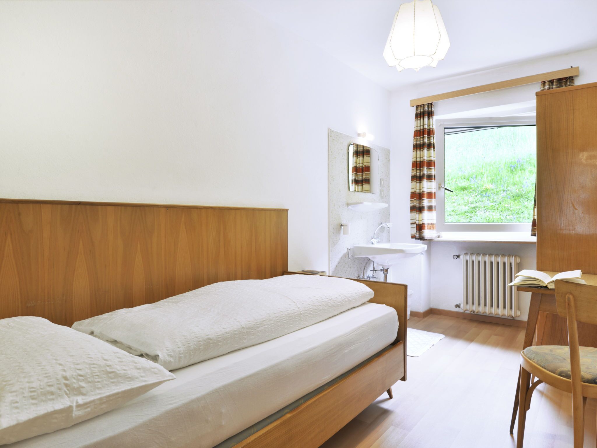 Ein Schlafzimmer mit einem hölzernen Bett, einem Nachttisch, einem Stuhl, einem Fenster mit Vorhängen und einer Heizung.