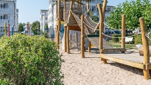 Een houten speelconstructie met een touwbrug en een houten platform, voor een achtergrond van witte gebouwen en een heldere lucht.