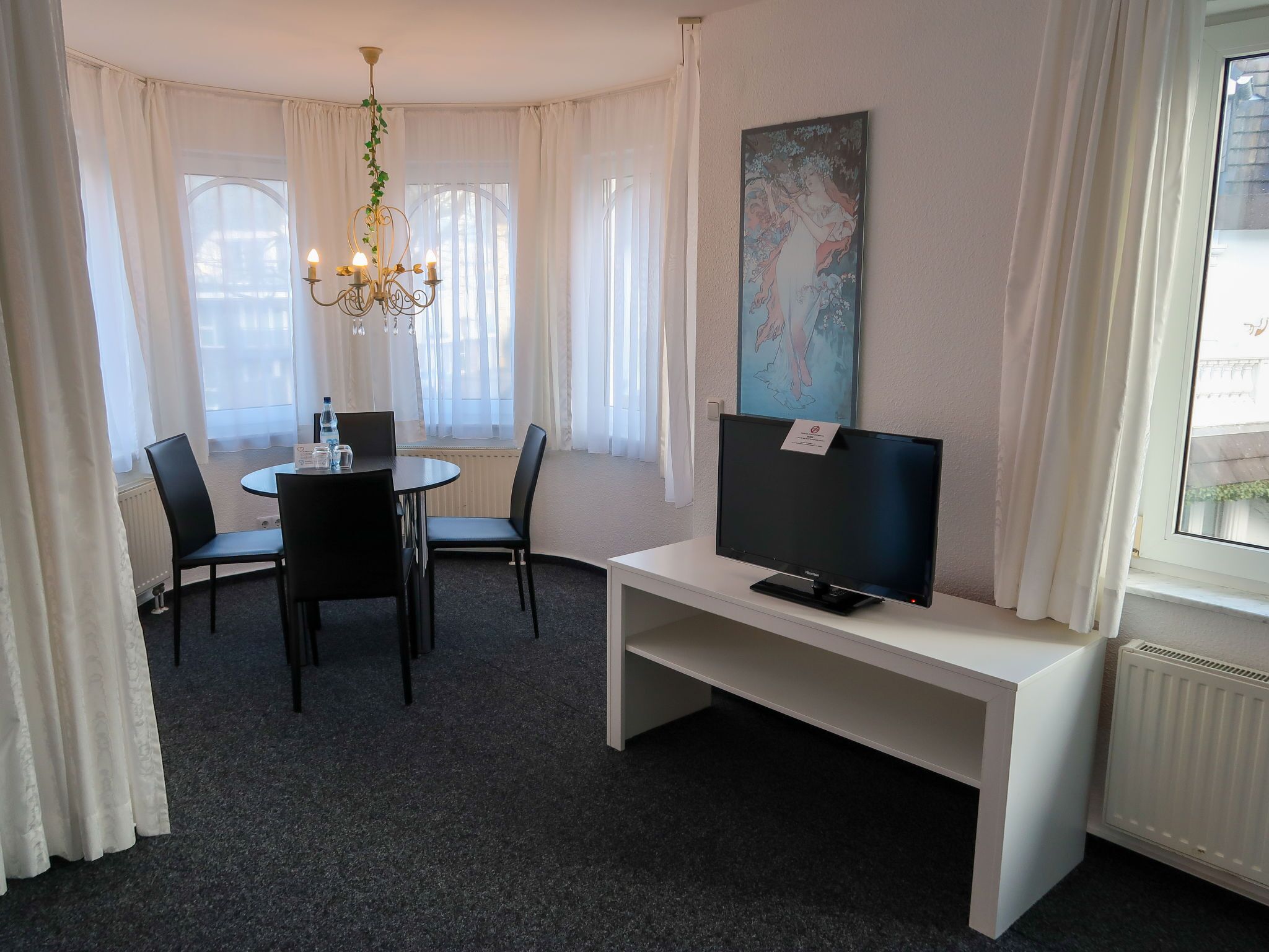 A hotel room with a dining table, chairs, a TV on a white stand, a chandelier, and a large window with curtains.