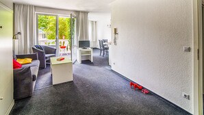 A modern hotel room with a sofa, a small table, a TV, and a balcony with a view of greenery. - Apartment Duhnen by Interhome (Cuxhaven)