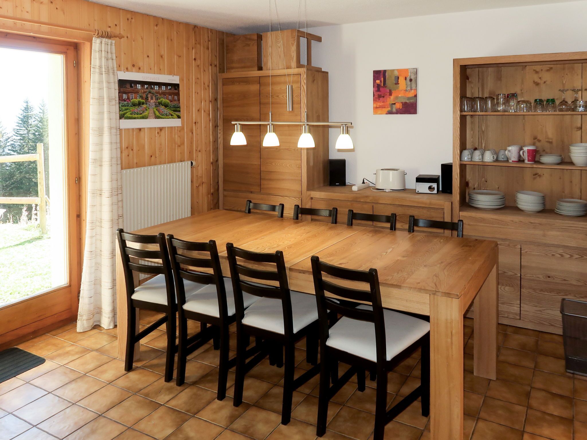 Ein Essbereich mit Holztisch und Stühlen, ein Küchenbereich mit Regalen und Blick ins Freie.