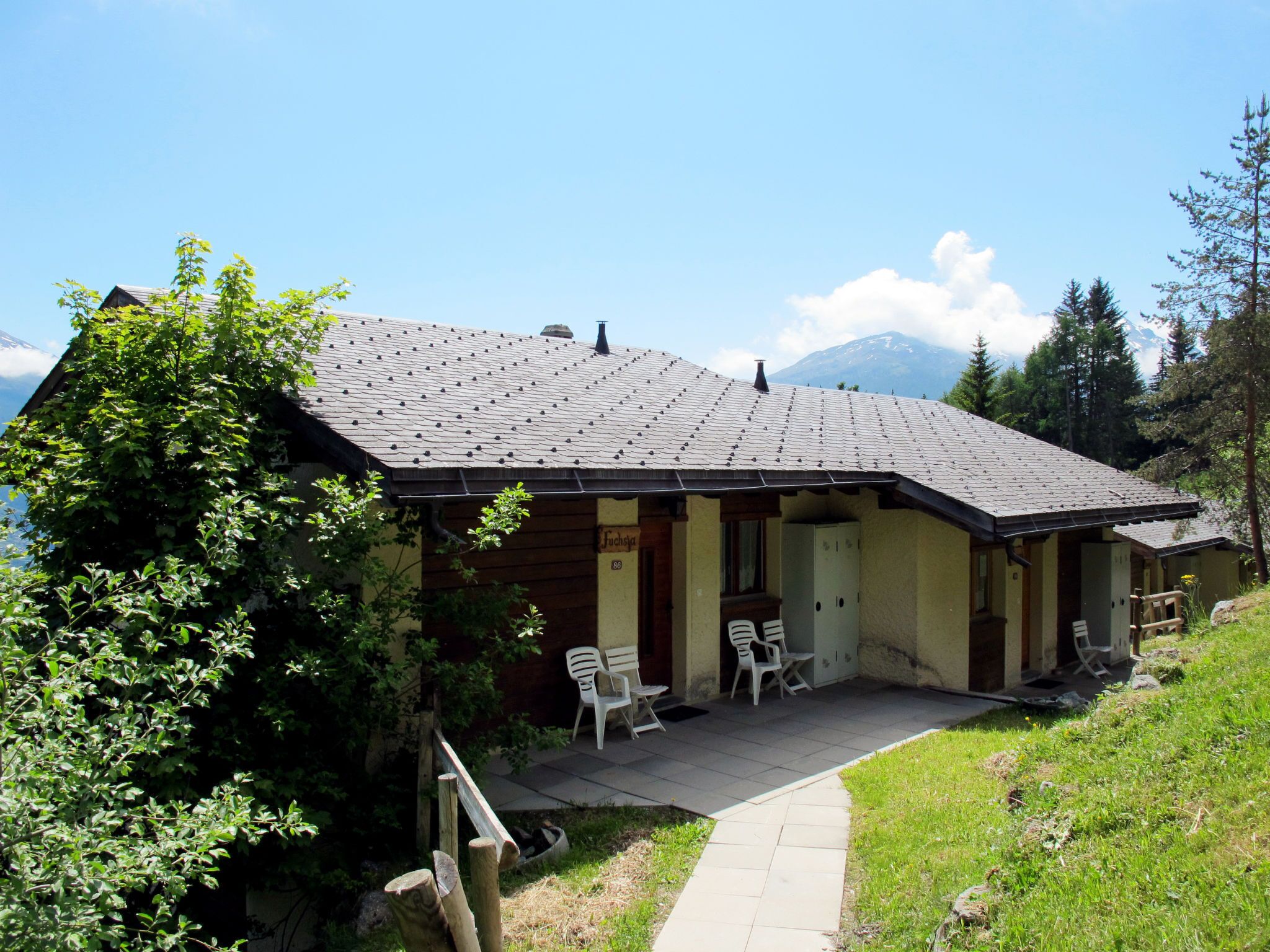 A chalet with a sloped roof, a covered porch with chairs, and a view of a mountain in the background.