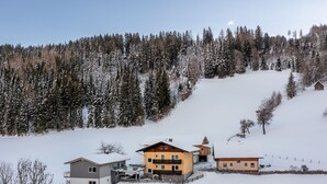 Un pueblo cubierto de nieve con varias edificaciones, incluyendo una casa amarilla destacada con techo inclinado, rodeado de bosques densos y una colina nevada.