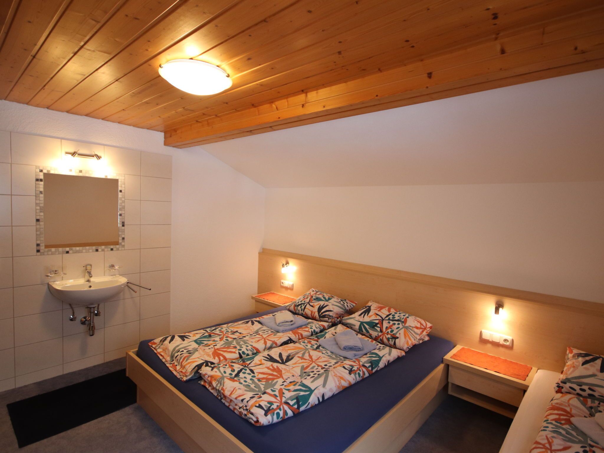 A bedroom with a wooden ceiling, a bed with a floral bedspread, a nightstand, and a wall-mounted mirror.