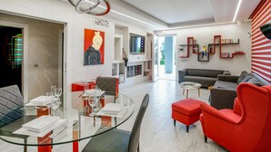 A modern living room with a glass dining table, red and grey sofas, and a TV mounted on the wall.