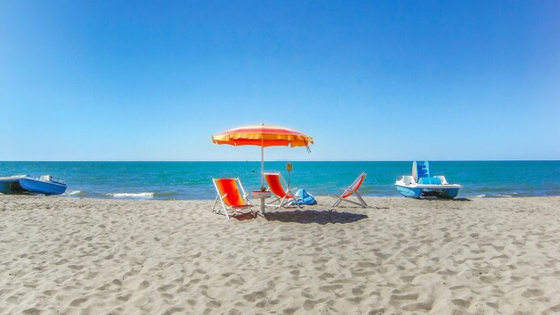 A beach with two lounge chairs under a red and yellow umbrella, a small boat, and a clear blue sky.