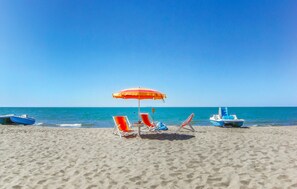 Une plage avec deux chaises longues sous un parasol rouge et jaune, un petit bateau et un ciel bleu clair.