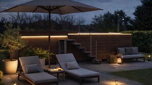 An outdoor seating area with lounge chairs and a large umbrella, a wooden deck, and a staircase leading to an upper level.