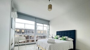 A modern bedroom with a large bed, a chair, and a view of buildings through large windows.