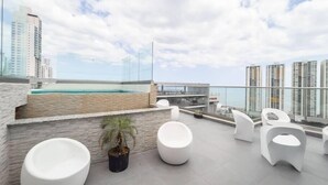 A rooftop terrace with a pool, white seating, and a view of high-rise buildings.
