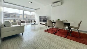 A modern living room with a sofa, dining table, chairs, a TV, and large windows.