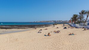 Una extensa playa de arena con gente tomando el sol y nadando, palmeras y un cielo azul despejado.
