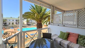 Un balcón con vista a una piscina y palmeras, una mesa redonda y un sofá con cojines.