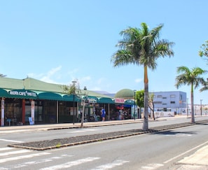 A street with shops including 'Andra Cafe Pastel' and 'China Shop'. Palm trees line the sidewalk. - Vis Herbae Pool View by iRent Fuerteventura (Corralejo)