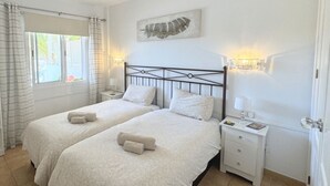 Un dormitorio con dos camas, una ventana con cortinas, una obra de arte en la pared, lámparas de noche y una mesita de noche con un libro y un vaso.