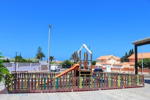 A playground with a slide and climbing structures, surrounded by a fence with colorful posts. - Vis Herbae Pool View by iRent Fuerteventura (Corralejo)