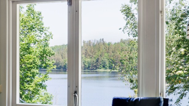 Ein Zimmer mit großem Fenster und Blick auf einen von Bäumen umgebenen See.